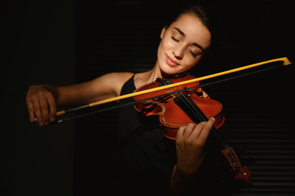 Beautiful young woman playing violin in dark room, focus on hand - Foto, afbeelding