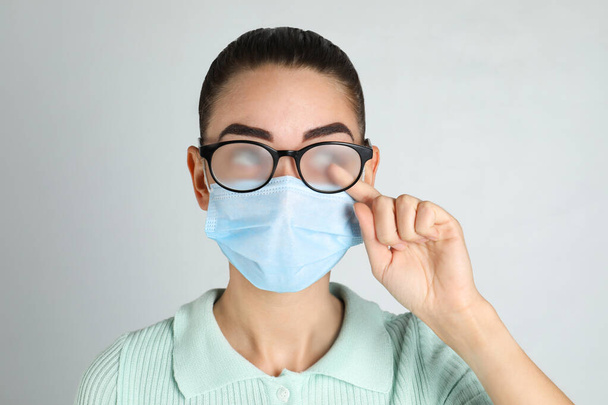 Woman wiping foggy glasses caused by wearing medical mask on light background - Φωτογραφία, εικόνα