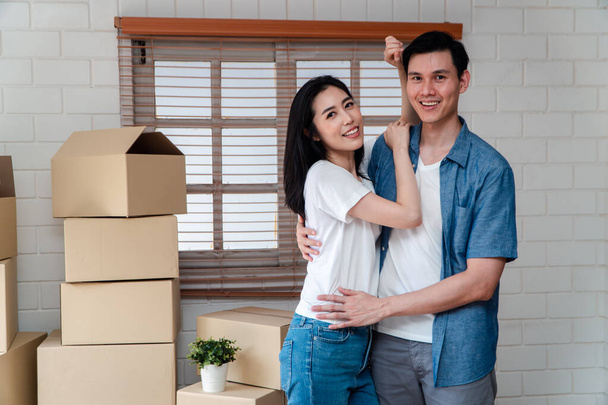 Улыбающаяся молодая азиатская счастливая пара обнимает парня картонными коробками во время переезда в новый дом после покупки недвижимости. Концепция начала новой жизни для молодоженов. - Фото, изображение