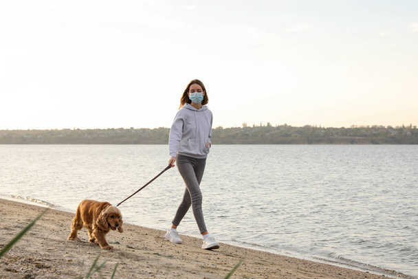 Woman in protective mask with English Cocker Spaniel on beach. Walking dog during COVID-19 pandemic - Фото, зображення