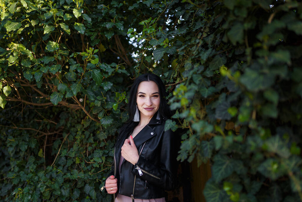 Portrait of young pretty dark-haired woman in black leather jacket posing on green ivy plant background. - Foto, immagini