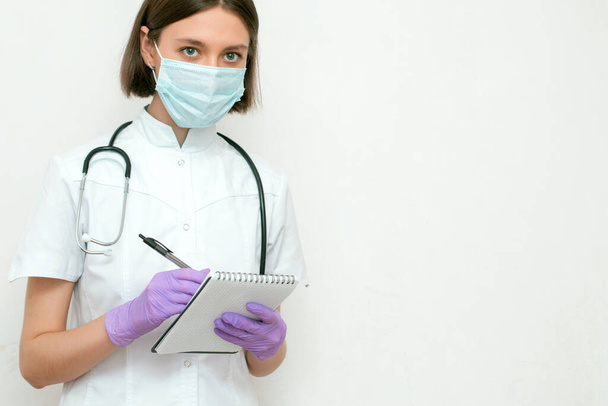 Nurse with stethoscope in white coat, gloves and medical mask holding clipboard, writing record and diagnosis on white background, free space. - Photo, image