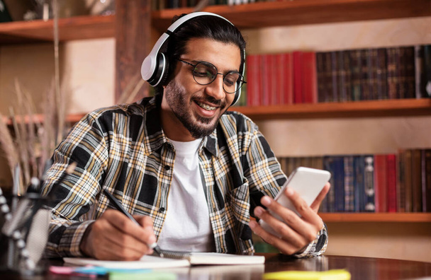 Ευτυχισμένος Ινδός φοιτητής με Smartphone Learning κάθεται στη βιβλιοθήκη του Πανεπιστημίου - Φωτογραφία, εικόνα