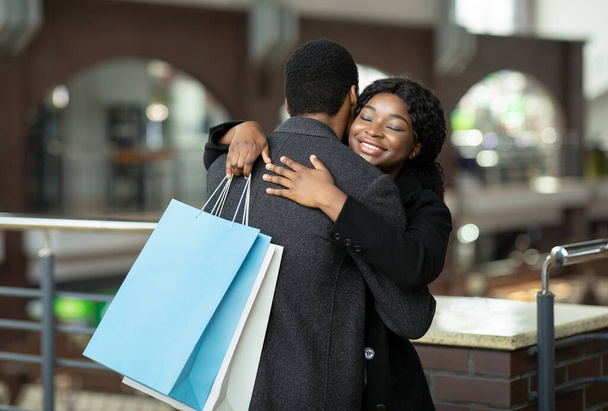 Szczęśliwy młody Afrykanin amerykański mężczyzna i pani w płaszczach przytula się w centrum handlowym podczas sezonowej sprzedaży i Czarny Piątek - Zdjęcie, obraz