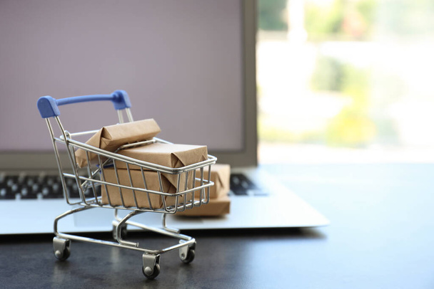 Internet shopping. Small cart with boxes near modern laptop on table indoors, space for text - 写真・画像