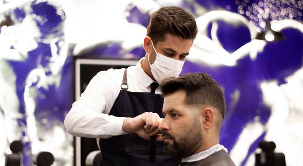 jeune et élégant coiffeur avec masque coupe les cheveux d'un client avec des caractéristiques caucasiennes - Photo, image