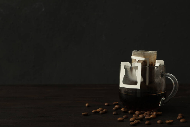 Glass cup with drip coffee bag and beans on black wooden table. Space for text - Fotografie, Obrázek