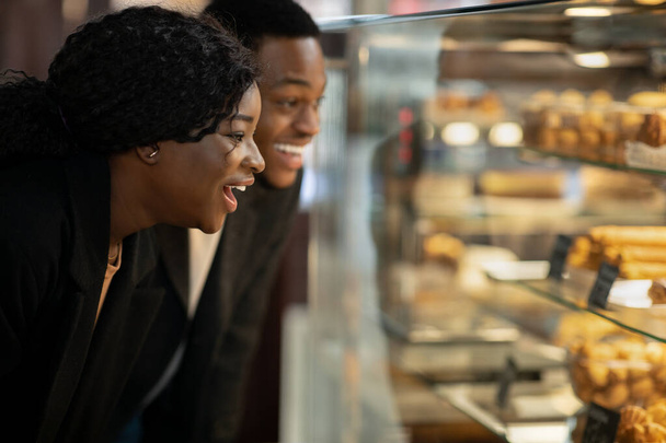 Überraschte glückliche Millennials Frau und Mann stehen vor Glasvitrine mit Gebäck und Kuchen - Foto, Bild