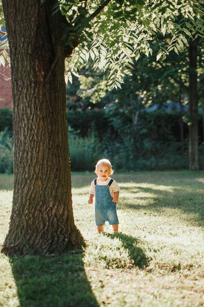 Lindo niño de pie bajo un enorme árbol grande en el parque al atardecer. Adorable niño sonriente de un año de edad niño caminando al aire libre en el patio trasero. Chico gracioso. Estilo de vida auténtico infancia feliz.  - Foto, imagen