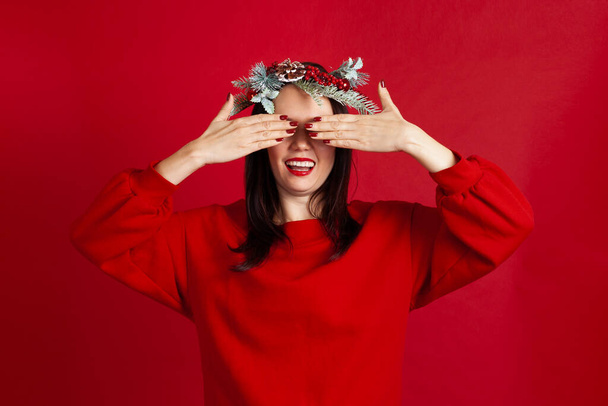 close-up portrait of surprised and excited Asian young woman in wreath covering her eyes with hands, on a red background - Foto, Imagen