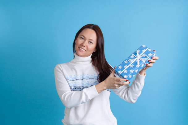 mock up smiling Asian young woman in sweater with snowflakes beautifully showing a gift box isolated on blue background - Φωτογραφία, εικόνα