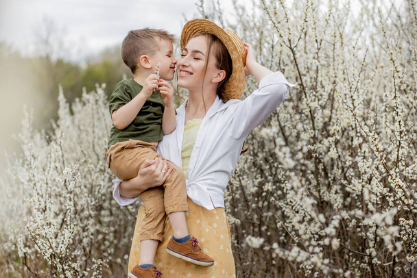 Glückliche Mutter und Sohn, die zusammen Spaß haben. Mutter umarmt ihren Sohn sanft. Im Hintergrund blühen weiße Blumen. Muttertag. - Foto, Bild