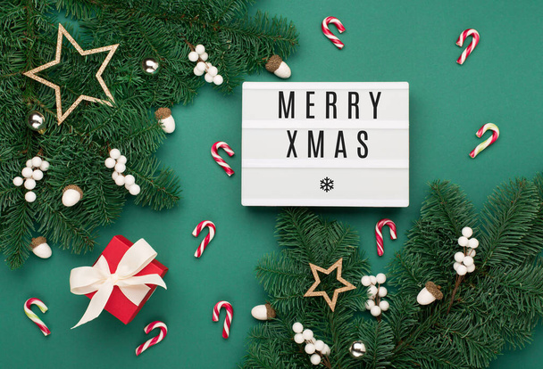 Merry Xmas text on white Lightbox with gift box, holiday gold decorations, acorns on green Christmas tree branches background. Flat lay, top view, copy space. Festive winter time. - Φωτογραφία, εικόνα