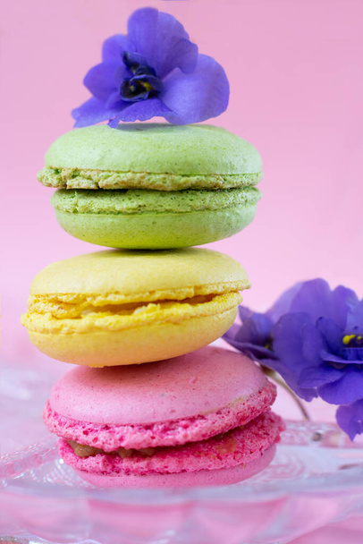 Pile of macarons decorated with african violet flowers on transparent glass plate.Vertical photo of french almond biscuits - Foto, Imagem