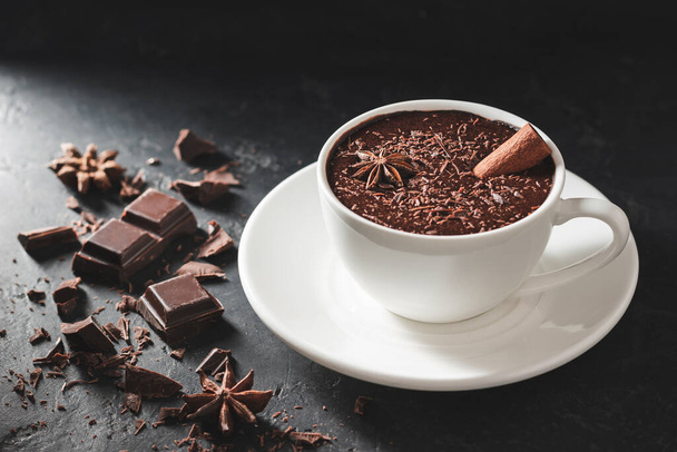 Hot chocolate drink in a white cup with cinnamon stick, broken chocolate cubes and star anise on textured dark background. - Photo, Image