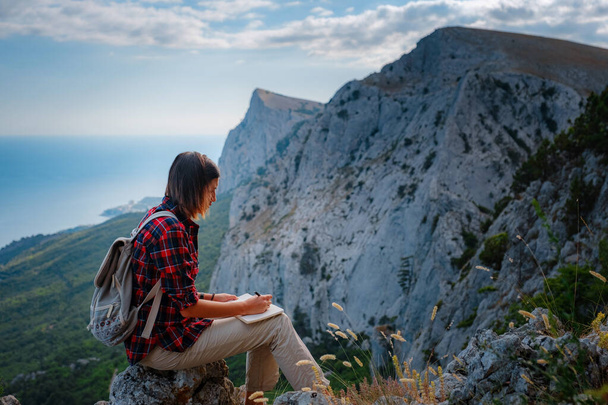vrouw wandelaar genieten van het uitzicht op bergtop klif, schrijft notities, ideeën in zijn papieren notitieboekje. idee van ecotoerisme reizen. Ontdekking Reisbestemming Concept - Foto, afbeelding