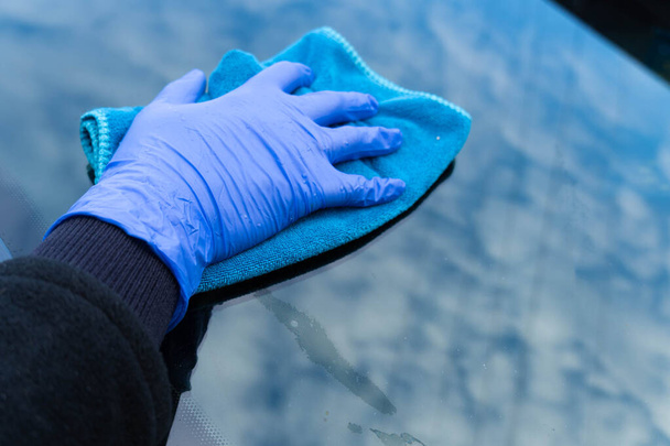 Naisen käsi sinisessä hanskassa hieroo auton lasia, jossa pilvet heijastuvat. Auton puhdistus - Valokuva, kuva