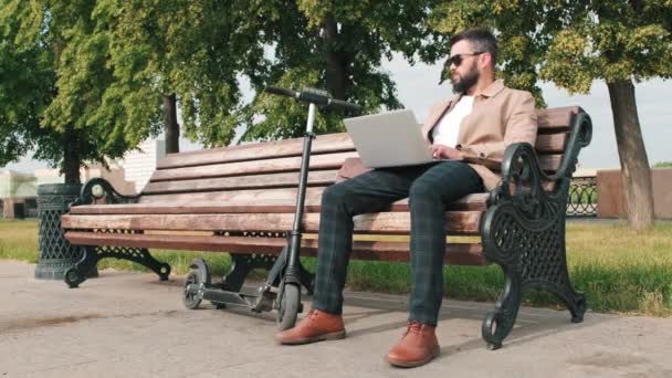 Volledig shot van moderne zakenman zittend op bank in park met elektrische scooter werkend op laptop en kijkend rond in daglicht - Video