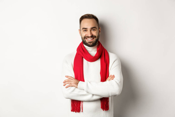 Vacanze di Natale e concetto di celebrazione. Bello uomo barbuto in maglione che celebra il nuovo anno, indossa sciarpa rossa e sorridente, in piedi su sfondo bianco - Foto, immagini