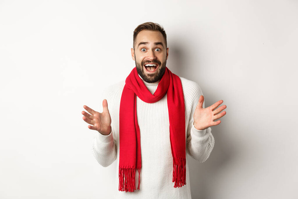 Weihnachtsfeiertage und Festkonzept. Bärtiger Typ schaut überrascht auf Neujahrspromo-Angebote, keucht erstaunt, trägt roten Schal und Pullover, weißer Hintergrund - Foto, Bild