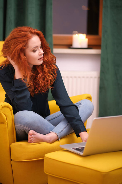 Nice beautiful lady with redhead curly hair work at the notebook sit down on the sofa at home - check on oline shops for cyber monday sales - technology woman concept for alternative office freelance - Fotografie, Obrázek