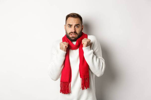 Рождественские праздники и концепция праздника. Испуганный мужчина дрожит от страха и смотрит испуганно, стоя взволнованный в красном шарфе и белом свитере - Фото, изображение