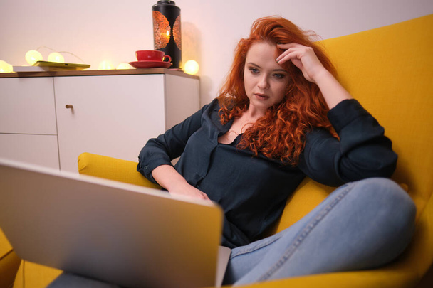 Nice beautiful lady with redhead curly hair work at the notebook sit down on the sofa at home - check on oline shops for cyber monday sales - technology woman concept for alternative office freelance - Fotoğraf, Görsel