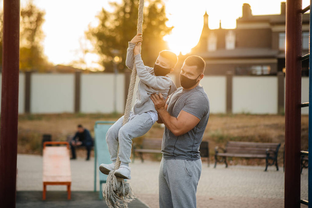 A father helps his son climb a rope on a sports field in masks during sunset. Healthy parenting and healthy lifestyle - Foto, Imagen