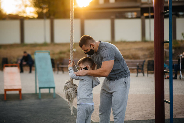 A father helps his son climb a rope on a sports field in masks during sunset. Healthy parenting and healthy lifestyle - Photo, image