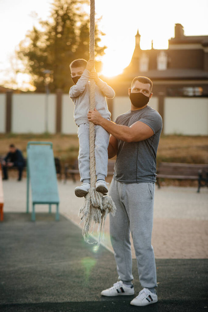 A father helps his son climb a rope on a sports field in masks during sunset. Healthy parenting and healthy lifestyle - Фото, зображення