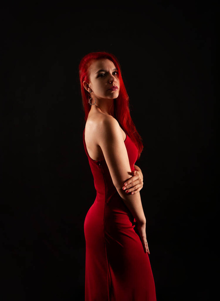 A girl with red hair, in a red dress posing on a dark background. Studio shooting . - Foto, afbeelding