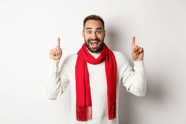 Рождественские покупки и зимние праздники концепция. Веселый красивый мужчина, показывающий рекламный баннер, указывающий пальцем на пространство для копирования, одетый в красный шарф и свитер, белый фон - Фото, изображение