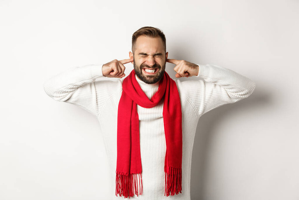 Winter holidays and shopping concept. Annoyed man shut his ears with fingers, distressed by loud noise or neighbours, standing against white background - Foto, Bild