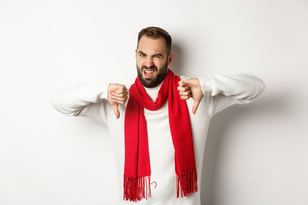 Skeptical and displeased man showing thumbs down, wearing winter sweater with scarf, dislike an disagree, standing over white background - Φωτογραφία, εικόνα