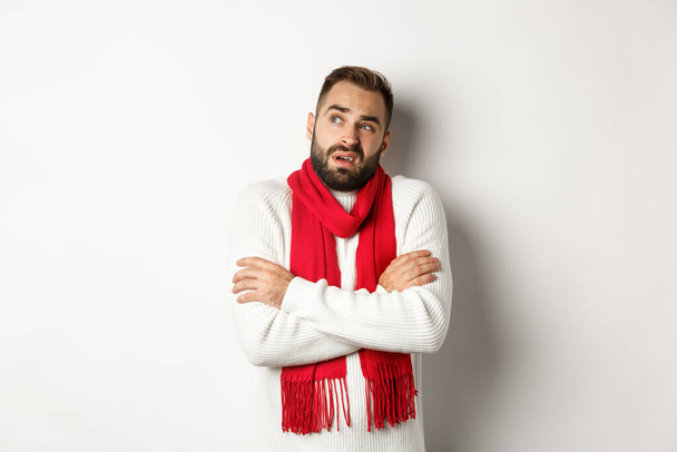 Homme confus regardant douteux, debout avec les mains croisées et se sentant froid en hiver, portant un pull de Noël avec une écharpe rouge, debout sur fond blanc - Photo, image