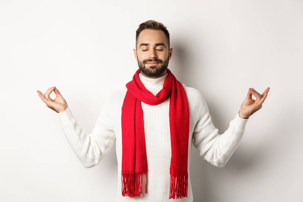 Ragazzo barbuto rilassato in piedi in pace, meditando con gli occhi chiusi, in piedi su sfondo bianco in sciarpa rossa e maglione - Foto, immagini