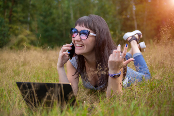 молода жінка лежить на траві з ноутбуком і емоційно говорить по телефону
. - Фото, зображення