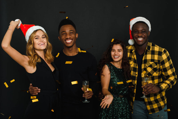 Farklı milletlerden dört arkadaş Noel Baba şapkalarına ve boynuzlarına konfeti atıp siyah arka planda şampanya içiyorlar. Gençlerden oluşan bir şirket Noel ve Yeni Yılı kutluyor - Fotoğraf, Görsel