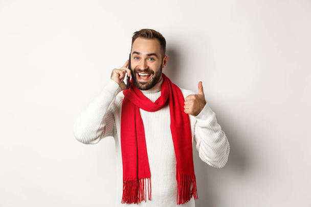 Handsome bearded man talking on phone, showing thumbs up in approval, like something, confirm order, standing over white background - Photo, Image