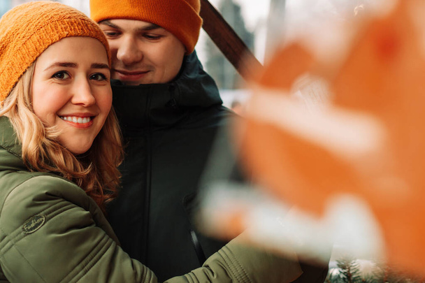 Mladý šťastný pár se spolu baví na vánočním trhu. Atraktivní muž a žena v oranžových kloboucích a zelených bundách objímají a líbají na ulici na Silvestra. Koncept zimní nálady - Fotografie, Obrázek