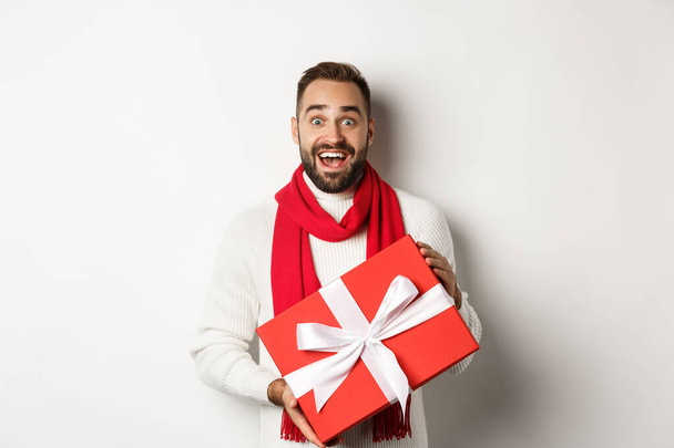 Concepto de vacaciones de invierno. Hombre sorprendido recibiendo regalo de Navidad y mirando sorprendido, sosteniendo caja envuelta roja con presente, de pie sorprendido en el fondo blanco - Foto, imagen