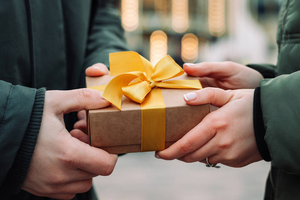 Hände eines Ehepaares mit einem Geschenk. Mann überreicht seiner Freundin auf der Straße ein kleines Geschenk mit gelbem Band. Frau nimmt Geschenk von ihrem Freund an. Valentinstag und Weihnachtskonzept - Foto, Bild