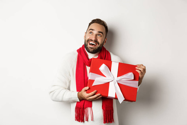 Καλά Χριστούγεννα. Ευτυχισμένος άνθρωπος που κοιτάζει εκστασιασμένος και κρατώντας το δώρο του για την Πρωτοχρονιά, στέκεται απέναντι σε λευκό φόντο - Φωτογραφία, εικόνα