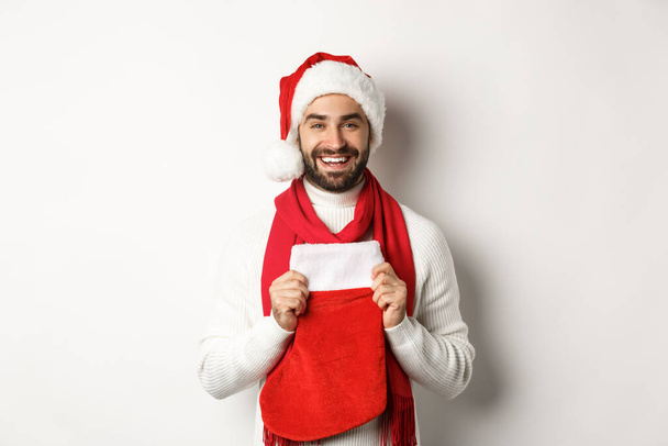 Zimowe wakacje i koncepcja zakupów. Przystojny brodaty mężczyzna przynieść prezenty w Boże Narodzenie skarpetki, uśmiechając się i życząc szczęśliwego Nowego Roku, białe tło - Zdjęcie, obraz