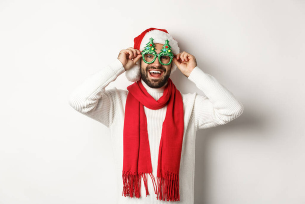 Χριστούγεννα, Πρωτοχρονιά και εορταστική ιδέα. Ευτυχισμένος άνθρωπος που γελάει, φοράει καπέλο και γυαλιά για πάρτι, στέκεται πάνω από λευκό φόντο - Φωτογραφία, εικόνα