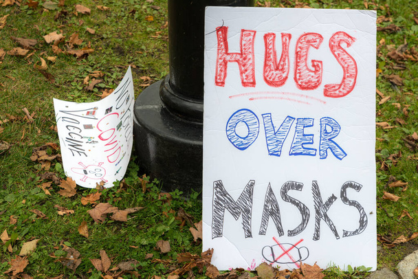 Μια αγκαλιά πάνω από την πινακίδα μάσκες. Σχεδιασμένη μάσκα με Χ στο κάτω μέρος. Ένα άλλο σημάδι διαμαρτυρίας για τα εμβόλια covid προς τα αριστερά, αλλά μόνο εν μέρει αναγνώσιμο. - Φωτογραφία, εικόνα