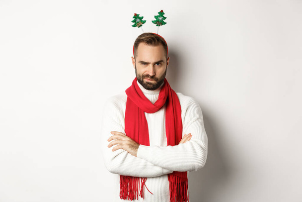 Christmas party and celebration concept. Suspicious young bearded man looking doubtful, wearing funny accessory hat, standing against white background - Fotoğraf, Görsel