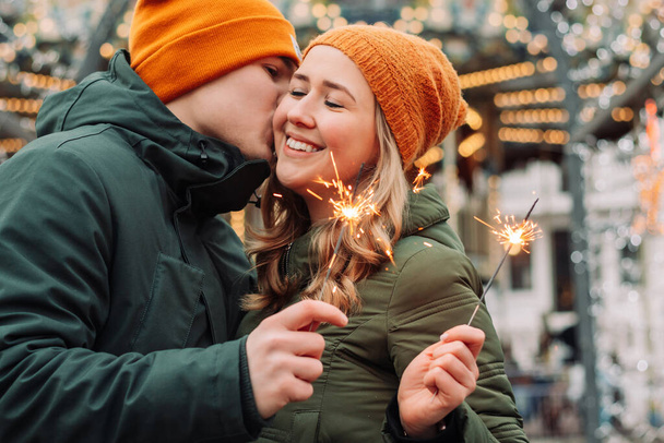 Young attractive couple hug and kiss outdoors with the sparklers in their hands celebrating New Year and Christmas. Winter holidays festival mood. Man and woman feel happy. Love and happiness concept - Foto, Imagem