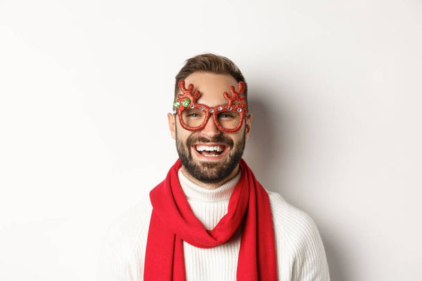 Christmas, New Year and celebration concept. Close-up of happy bearded man smiling, wearing party glasses and red scarf, white background - Photo, Image