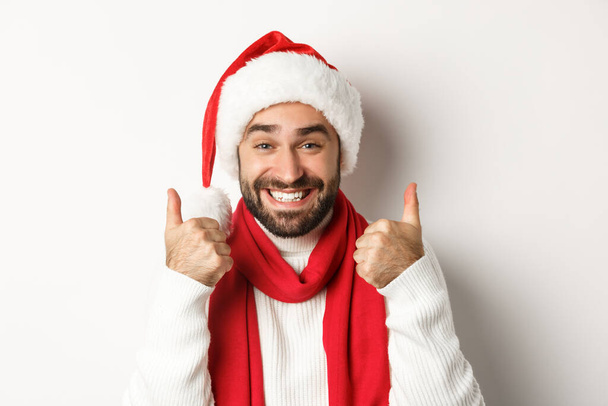 New Year party and winter holidays concept. Close-up of happy man in Santa hat showing thumbs up in approval, like and agree, standing over white background - 写真・画像